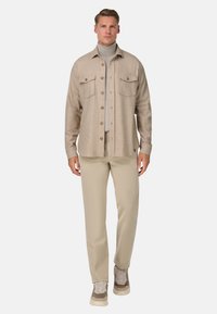 Béžová košile s knoflíky a dvěma náprsními kapsami, zkombinovaná s světle béžovými kalhotami a teniskami s kombinací hladkých a texturovaných materiálů.