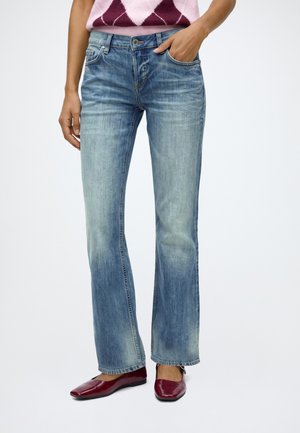 Personne portant un jean bleu délavé à jambes droites, un pull à losanges rose et bordeaux, une main dans la poche et des chaussures plates rouges.