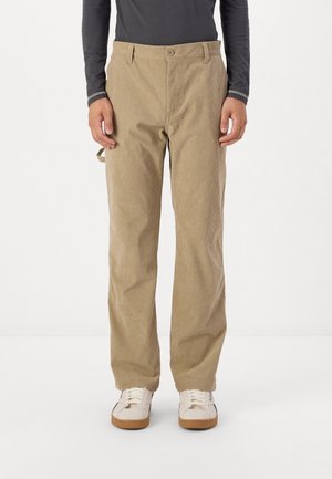 Beige corduroy trousers with a straight leg, featuring a button closure and side pockets. Worn with white sneakers and a dark long-sleeve shirt.