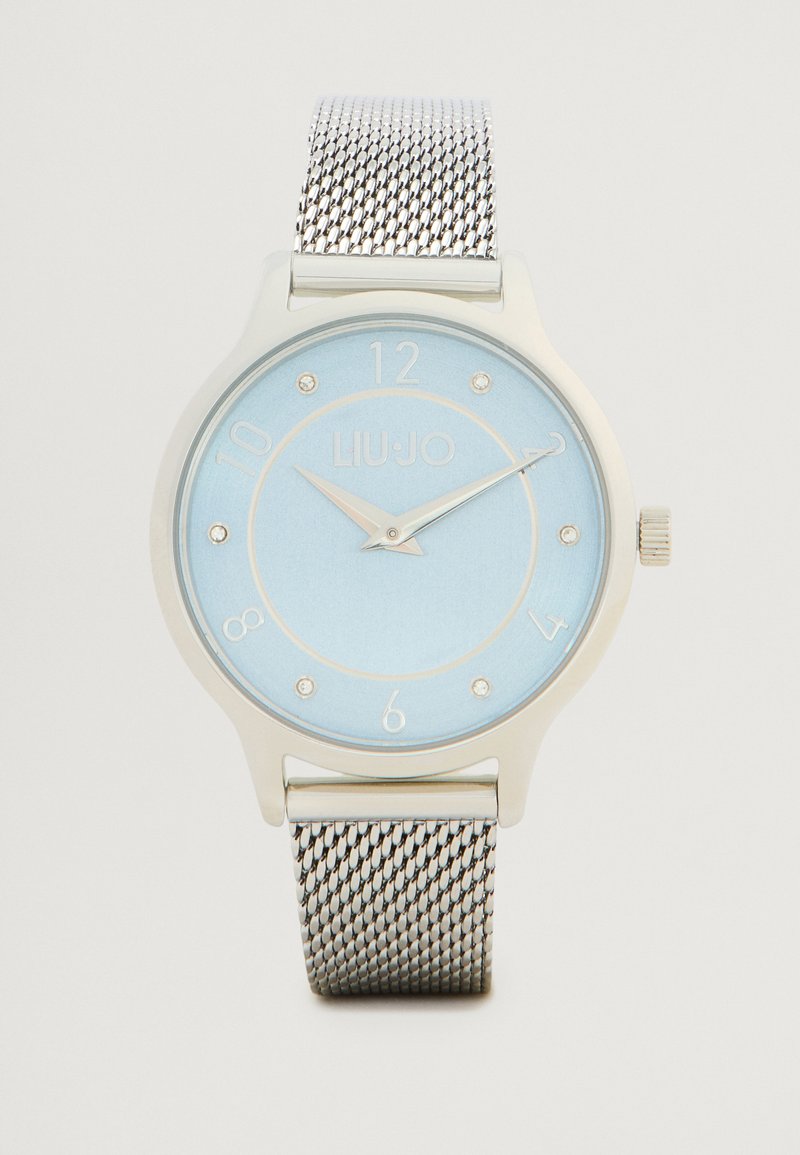 Ρολόι με ασημένια πλέξη διορθωτή με ανοιχτό μπλε καντράν, που διαθέτει ασημί δείκτες ωρών και λογότυπο. Στρογγυλή κάσα με λεία επιφάνεια και κορώνα.