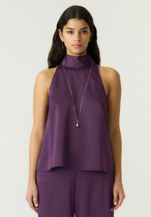 Femme aux longs cheveux foncés portant un haut violet sans manches et un pantalon assorti, accessoirisée d’un long collier argenté avec un pendentif.