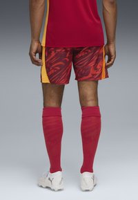 Czerwony i żółty strój sportowy z wzorzystymi szortami, czerwonymi skarpetami do kolan i białymi korkami z szarymi akcentami. Tylny widok ukazuje szczegóły projektu.