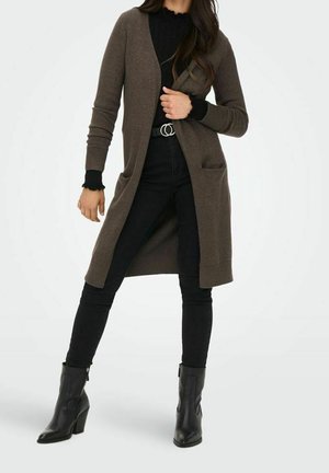 Frau, die eine lange braune Strickjacke über einem schwarzen Oberteil trägt, schwarze Jeans mit einem Doppelring-Gürtel und schwarze Stiefeletten, steht mit gespreizten Beinen.