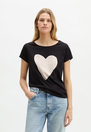 Kobieta o długich blond włosach, ubrana w czarny t-shirt z dużym białym sercem i jasnoniebieskie dżinsy, stojąca z jedną ręką w kieszeni.