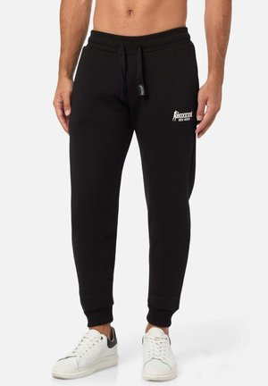 Schwarze Baumwoll-Sweatpants mit einem Kordelzugbund, schmal zulaufenden Beinen und einem weißen Logo auf dem linken Oberschenkel. Kombiniert mit weißen Sneakern.