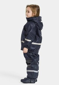 Marinblå vattentät regnoverall med reflexband, huva, elastiska mudd och mönstrade förstärkningar på knäna. Svarta gummistövlar.