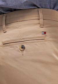 Beige pants pocket with button and small red, white, and blue logo detail, worn with tucked-in blue shirt.