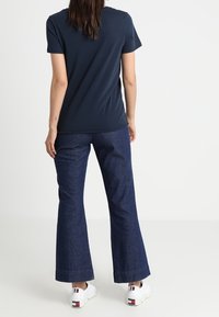 Žena stojící s otočením zády, oblečená v tmavě modrém tričku, tmavě modrých rozšířených džínách a bílých teniskách s červenými a modrými detaily.