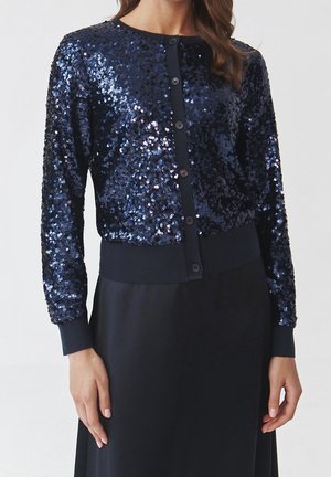 Vrouw draagt een marineblauwe lange mouwen pailletten cardigan met zwarte knopen, gecombineerd met een zwarte rok tegen een effen achtergrond.