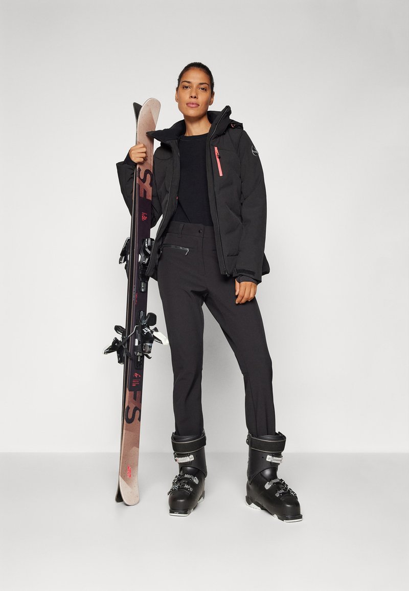 Schwarze gepolsterte Skijacke mit rosa Reißverschluss, schwarzer Pullover, schwarze Skihose und schwarze Skischuhe. Hält Skier mit schwarzen Bindungen.