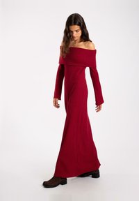 Robe rouge à épaules dénudées, à manches longues, avec une silhouette ajustée, une texture douce et une jupe fluide. Portée avec des bottines marron.