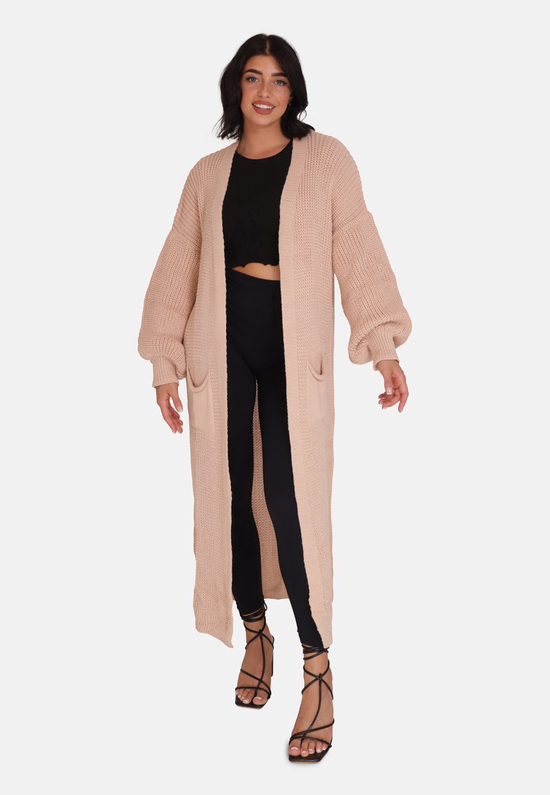 Lång, stickad cardigan i ljusbeige med ribbad textur, öppen front och två framfickor, kombinerad med svarta leggings och sandaler med remmar.