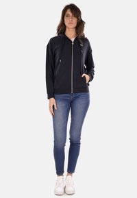 Felpa nera con zip e cappuccio, dotata di una cerniera dorata e tasche frontali. Abbinata a jeans skinny blu e sneakers bianche.