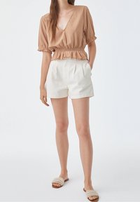 Blusa marrone con texture e vita elasticizzata, pantaloni corti bianchi plissettati e sandali a soffietto beige intrecciati. Un outfit semplice e casual in tonalità neutre.