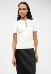 Biała bluzka z krótkim rękawem o dopasowanym kroju z okrągłym dekoltem, z dekoracyjnymi złotymi i przezroczystymi guzikami wzdłuż przodu.
