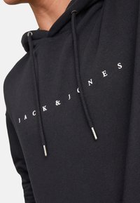 Sort hættejakke med snore. Har hvid tekst "JACK & JONES" hen over fronten. Materialet virker blødt med en glat tekstur.