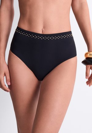 Bas de bikini noir taille haute avec de petites clous dorés le long de la ceinture, porté par une personne debout avec des bracelets visibles à un poignet.