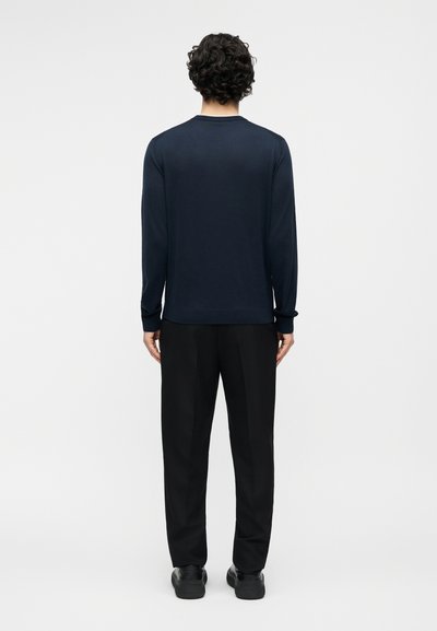 Homme aux cheveux bouclés vu de dos, portant une chemise bleu marine unie à manches longues, un pantalon noir et des chaussures noires, devant un fond blanc.