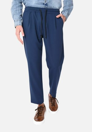 Uomo che indossa pantaloni con coulisse in vita elasticizzata blu navy, sneakers in pelle marrone e una camicia di jeans azzurro chiaro.