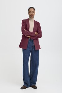 Burgundy blazer över en beigh turtleneck, kombinerat med vida mörkblå denimjeans och bruna skor, mot en enkel vit bakgrund.