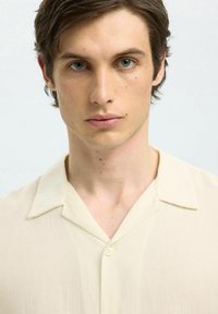 Camicetta corta beige chiaro con colletto a camicia, con una chiusura a bottoni sul davanti e un tessuto morbido e texturizzato.