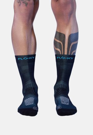 Benen met zwarte hoogpresterende sokken met label "FLY KY" en links- en rechtsindicatoren, die kuittattoos laten zien tegen een witte achtergrond.