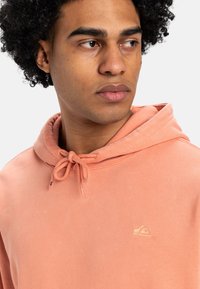 Sudadera de coral claro con capucha y cordón, textura suave y un pequeño logo dorado en el pecho. La capucha está en una tonalidad ligeramente más oscura.