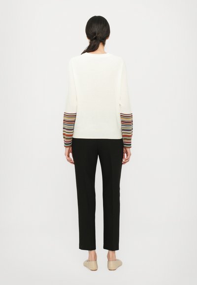 Maglione a maglia color crema con maniche a righe multicolori, abbinato a pantaloni neri e scarpe beige senza lacci. Tessuto liscio, vestibilità casual.