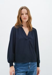 Donna che indossa una blusa blu navy a maniche lunghe con polsini arricciati e scollatura a V, abbinata a jeans blu, in piedi su uno sfondo bianco semplice.