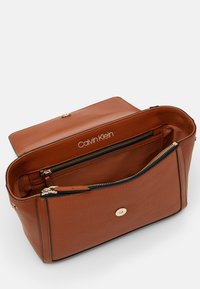 Hnědá texturová kožená kabelka se zipovou kapsou a zlatým kováním. Interiér ukazuje logo a funkční přihrádky.