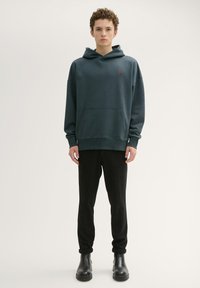 Przesadzony ciemnozielony kaptur z kieszenią kangurzą, żebrowanymi mankietami i małym czerwonym logo; w komplecie z czarnymi spodniami i czarnymi botkami za kostkę.