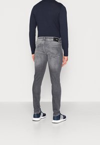 Man med åtsittande blekta grå jeans, marinblå långärmad tröja och marinblå sneakers med vita ränder, står vänd bort från kameran mot vit bakgrund.
