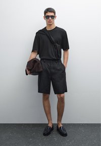 T-shirt nera a maniche corte, pantaloni corti neri su misura, mocassini neri, occhiali da sole grigi e una giacca marrone gettata su una spalla.