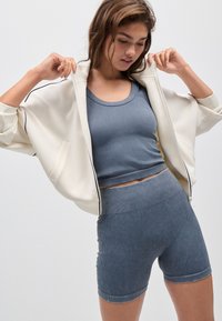 Lättviktskrämjacka med dragkedja över en figurnära, ribbad blå crop top och matchande shorts, med texturerat tyg och avslappnad passform.