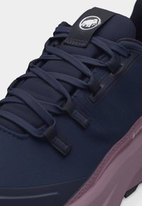 Artimas vaizdas tamsiai mėlynų sportinių batelių su storu rožiniu padu, detaliu raištelių surišimu ir baltu mamuto logotipu ant liežuvėlio juostelės.