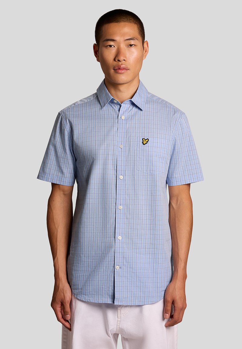 Mand iført lyseblå, kortærmet ternet skjorte med gult broderet logo på brystlommen, stående foran en ensfarvet grå baggrund.