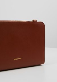 Braune Ledertasche mit glatter Textur, rechteckiger Form, goldenem Reißverschluss und geprägt "ROYAL REPUBLIQ"-Logo auf der Vorderseite.