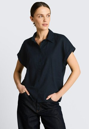 Frau trägt kurzärmliges marineblaues Hemd mit Knöpfen und dunkle Jeans, steht mit den Händen in den Taschen und schaut nach links vor neutralem Hintergrund.