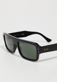 Ochelari de soare dreptunghiulari negri cu lentile verzi, având un design de ramă îndrăzneț și accente de logo aurii pe tâmple. Fără modele vizibile.