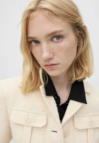 Chaqueta de tweed color crema con dos bolsillos de solapa y cierres de botón, combinada con una camisa negra con cuello debajo.