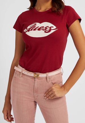 Vrouw draagt een rood t-shirt met het "Guess"-logo, ingestopt in een hoogwater roze jeans met een lichtroze riem met een gouden gesp.
