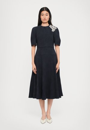Vestido navy con mangas cortas abullonadas, cuerpo ajustado, falda acampanada, tela texturizada y adornos florales en el hombro izquierdo.