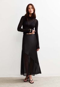 Haut noir à manches longues avec un tissu texturé, design court, et poignets sans doigts, associé à une jupe noire asymétrique fluide et des claquettes.