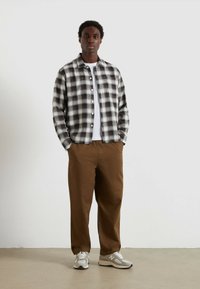 Zwart-wit geblokte flanellen shirt met knoopsluiting aan de voorkant, gecombineerd met tan kleurige broek. Model draagt grijze sneakers. Eenvoudige achtergrond.