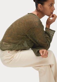 Pull en laine vert olive avec un design lâche et texturé, doté de manches larges et d'un col rond, porté avec un pantalon de couleur crème.