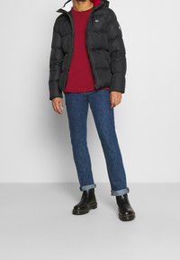 Zwarte pufferjack met een rood gevoerde capuchon, donkerrode t-shirt, blauwe jeans en zwarte enkellaarsjes. Jas heeft een ritsluiting en merkdecoratie.