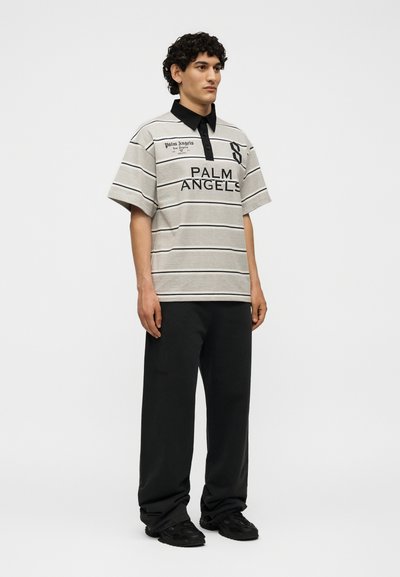 Άνδρας μοντέλο που φοράει γκρι και μαύρο ριγέ πόλο μπλουζάκι Palm Angels, μαύρο παντελόνι και μαύρα αθλητικά παπούτσια, στέκεται μπροστά σε απλό φόντο.