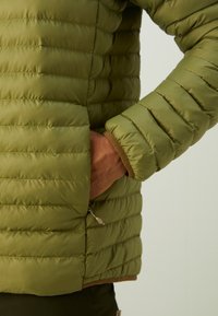 Olivenfarvet pufferjakke med en quiltet design. Har en sidelomme, en brun stofkant ved manchetterne og glat, skinnende materiale.