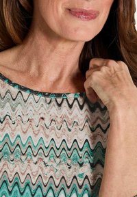 Gemusterte Bluse mit wellenförmigem Design in Teal, Schwarz und Creme. Verfügt über einen weiten Halsausschnitt und einen glatten, leichten Stoff. Nahaufnahme des Halsausschnitts und der Hand.