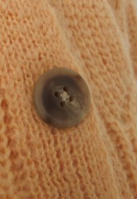 Närbild av persikofärgad stickad tyg med en cirkulär, något genomskinlig brun knapp som har fyra hål för fastsättning.